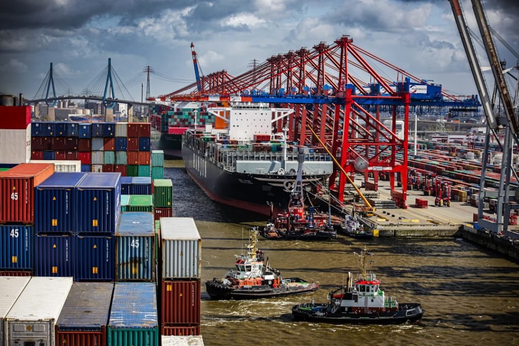 a-tug-boat-in-the-water-next-to-a-large-cargo-ship-hucilx-bvei Vibrant port scene with a large cargo ship being guided by tugboats, flanked by towering red cranes and stacks of multicolored shipping containers under a dynamic sky.
